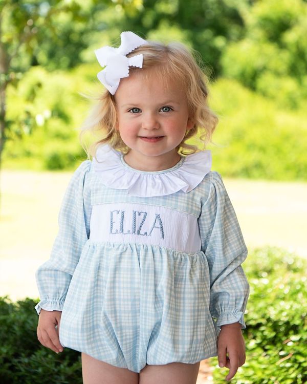 Young girl wearing a checkered dress with 'ELIZA' printed on it, standing outdoors.