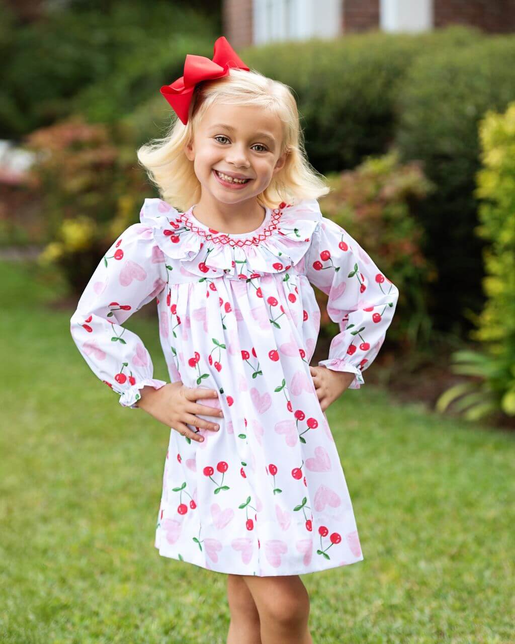Young girl wearing a cherry-patterned dress with a red bow outdoors.