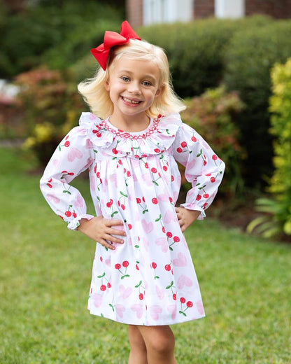 Young girl wearing a cherry-patterned dress with a red bow outdoors.