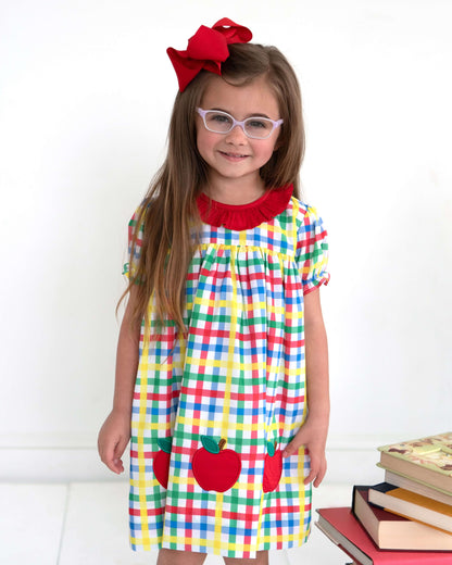 Young girl wearing a colorful dress with red apples and a red bow, standing against a white background.