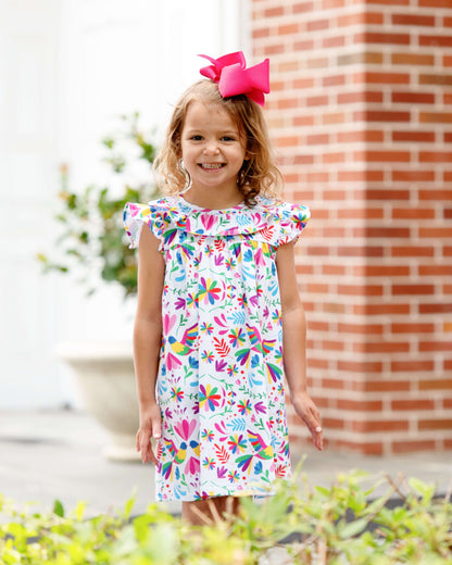 Young girl wearing a colorful floral dress with a pink bow, standing in front of a brick wall.