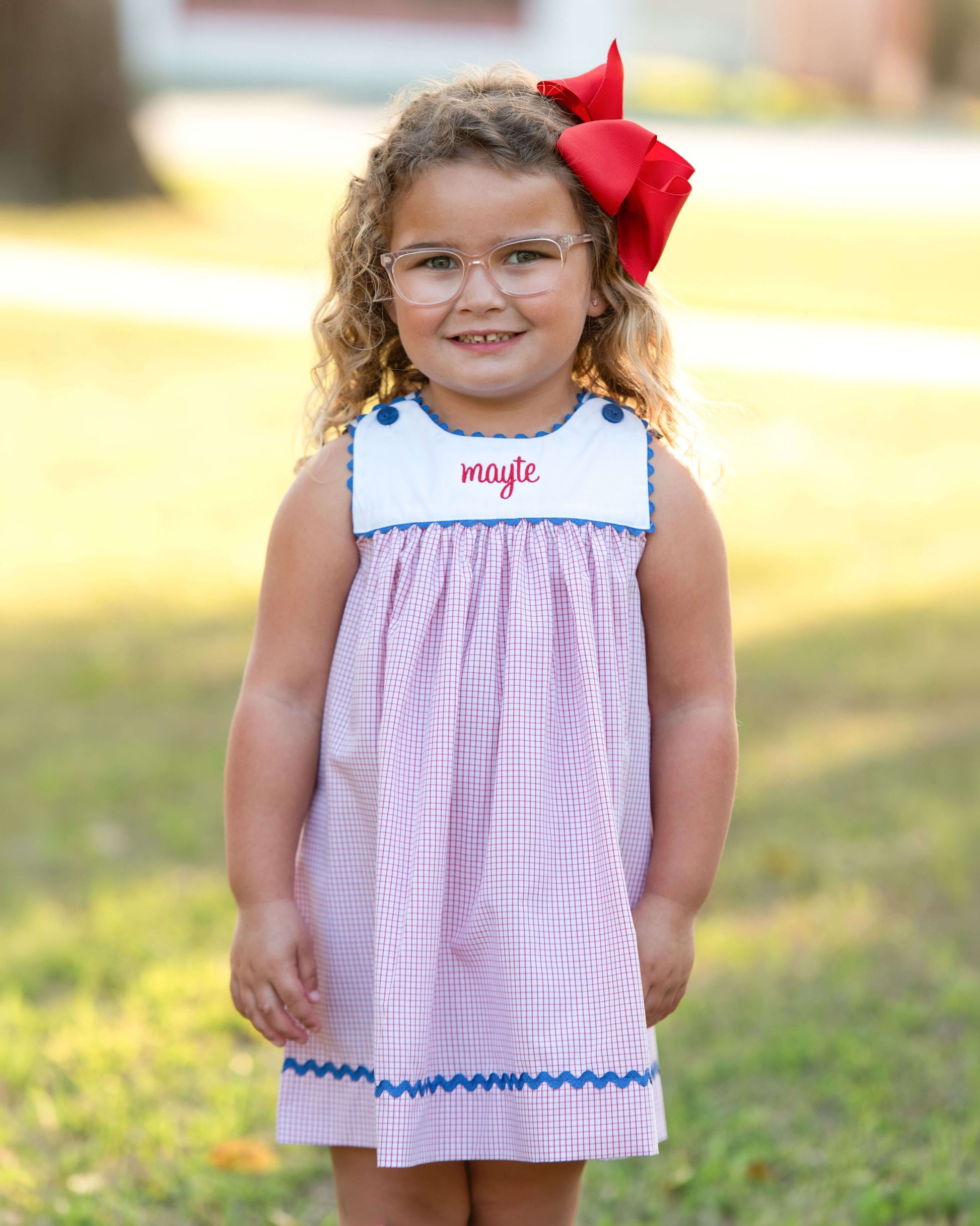 Young girl wearing a dress with 'maylee' embroidered on it, standing outdoors.