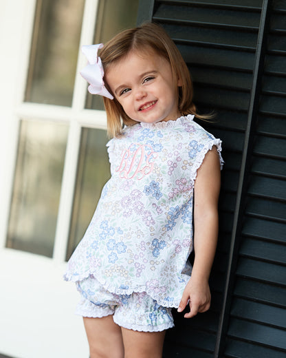 Young girl wearing a floral diaper set with a white bow in her hair, standing against a dark background.