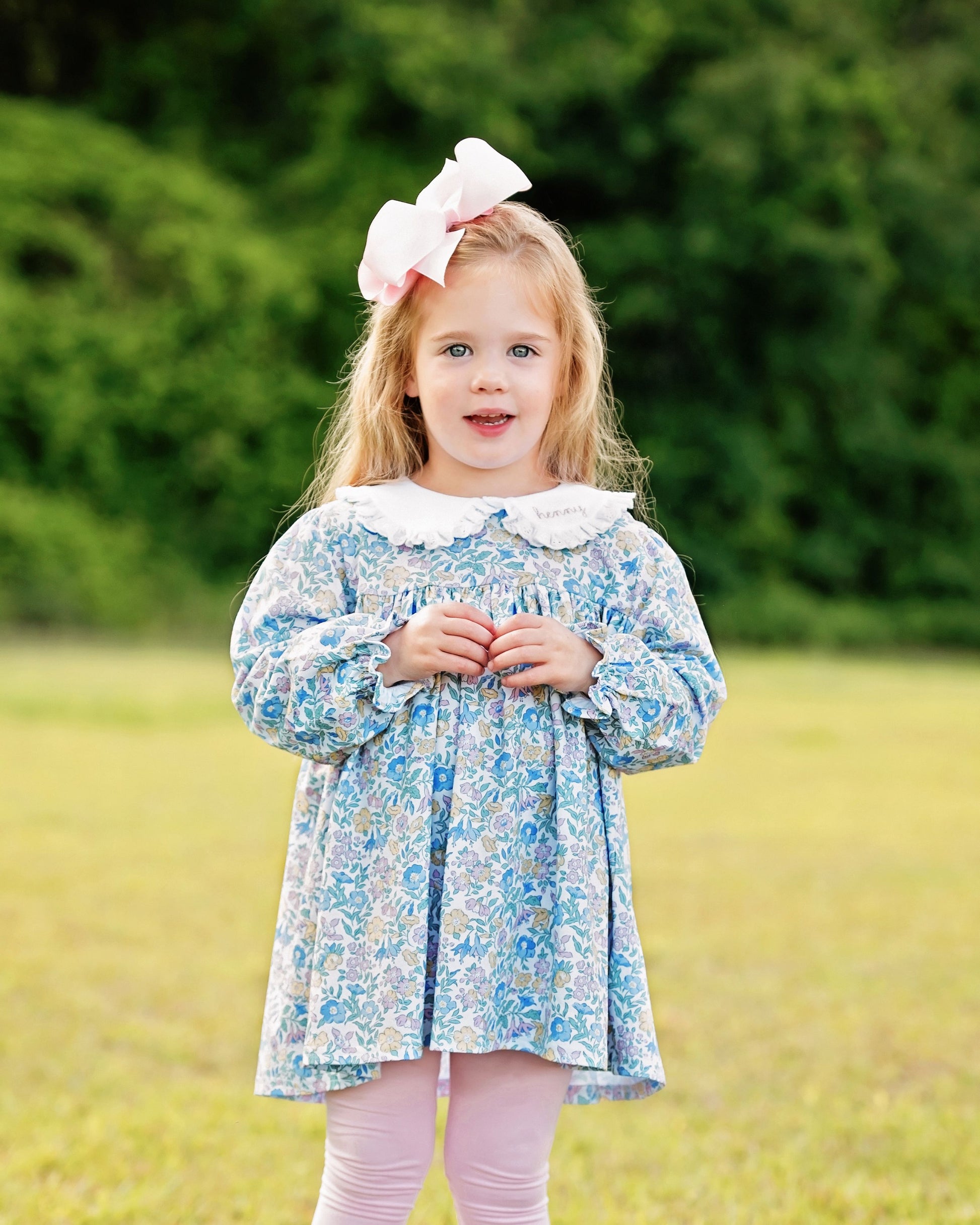 Young girl wearing a floral dress with a large bow in her hair, standing in a grassy field.