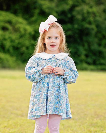 Young girl wearing a floral dress with a large bow in her hair, standing in a grassy field.