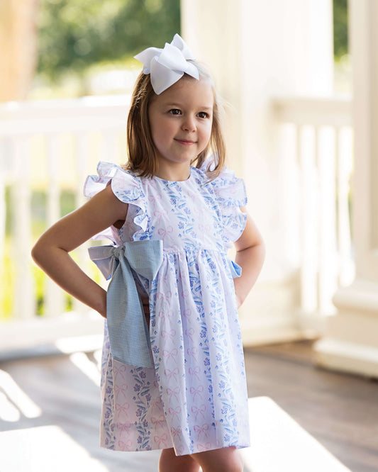 Young girl wearing a floral dress with a large bow, standing outdoors.