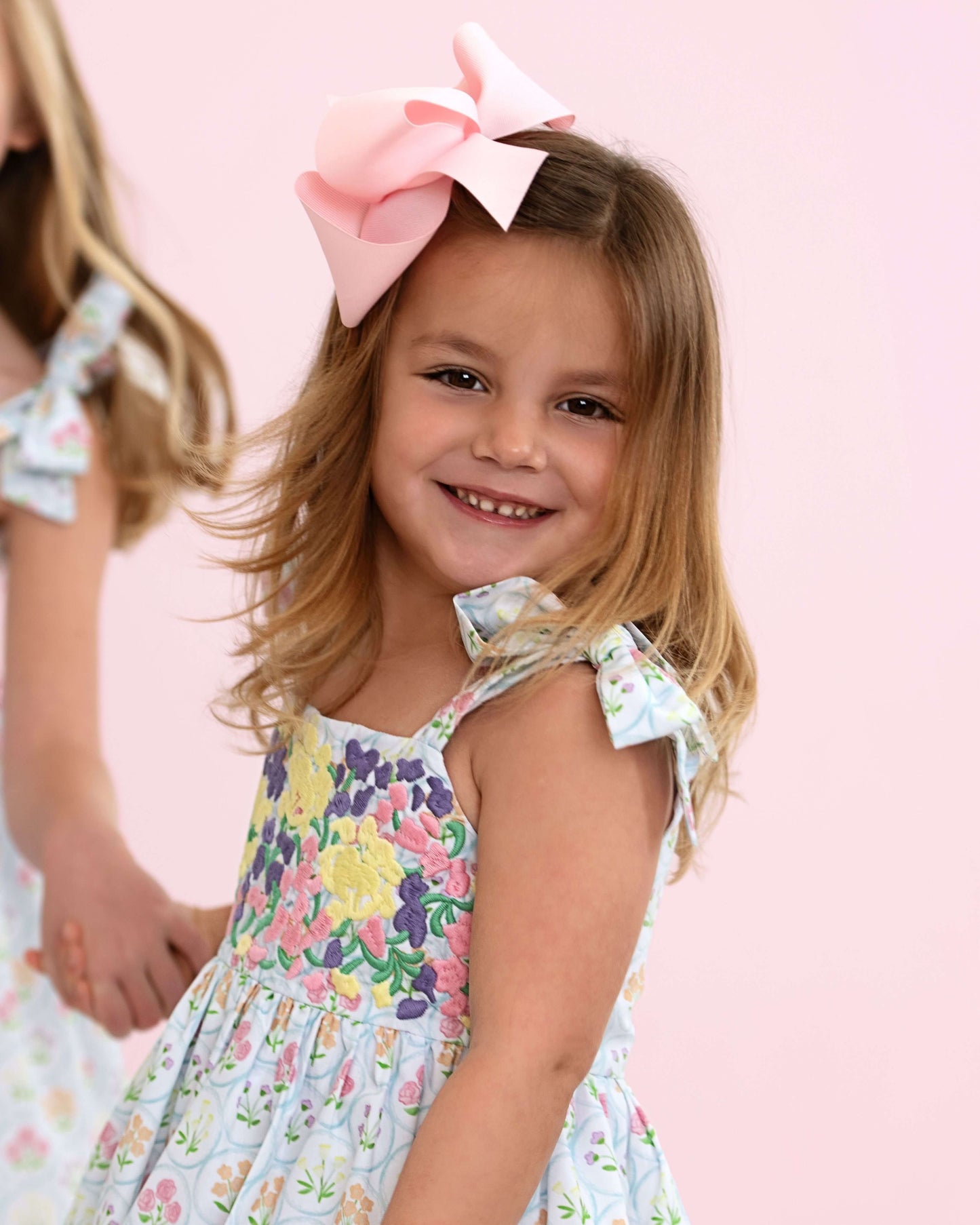 Young girl wearing a floral dress with a large pink bow in her hair against a pink background