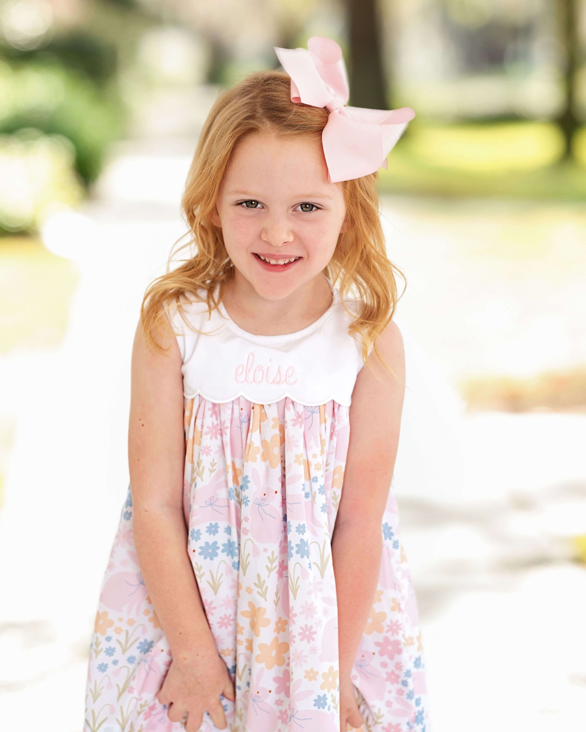 Young girl wearing a floral dress with a pink bow in her hair, standing outdoors.