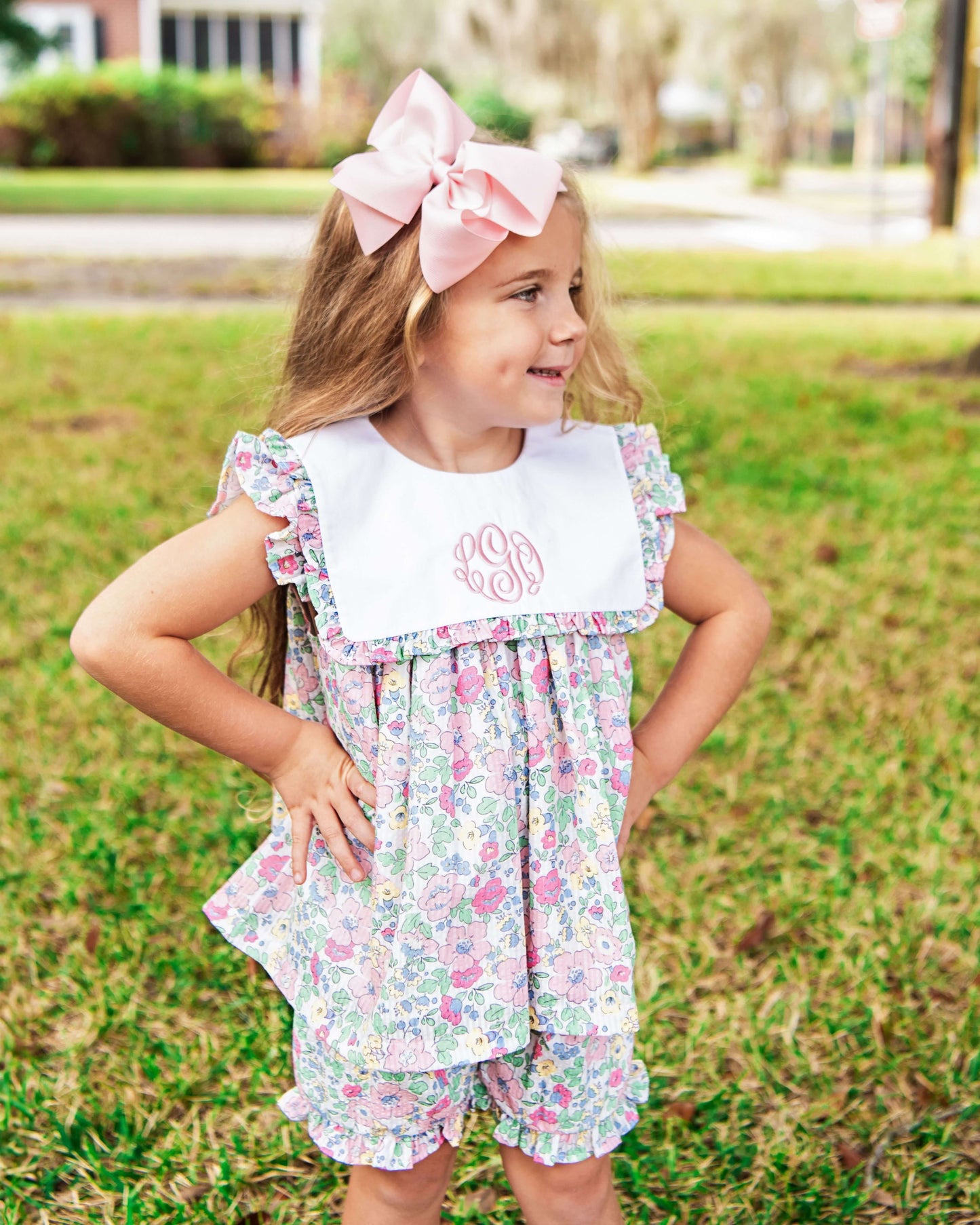 Young girl wearing a floral dress with a white bib and pink bow outdoors.