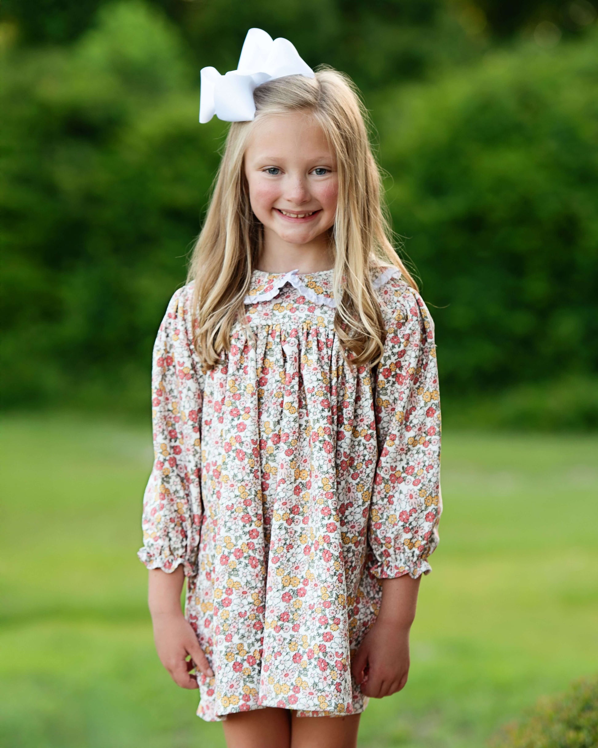 Young girl wearing a floral dress with a white bow in her hair, standing outdoors.