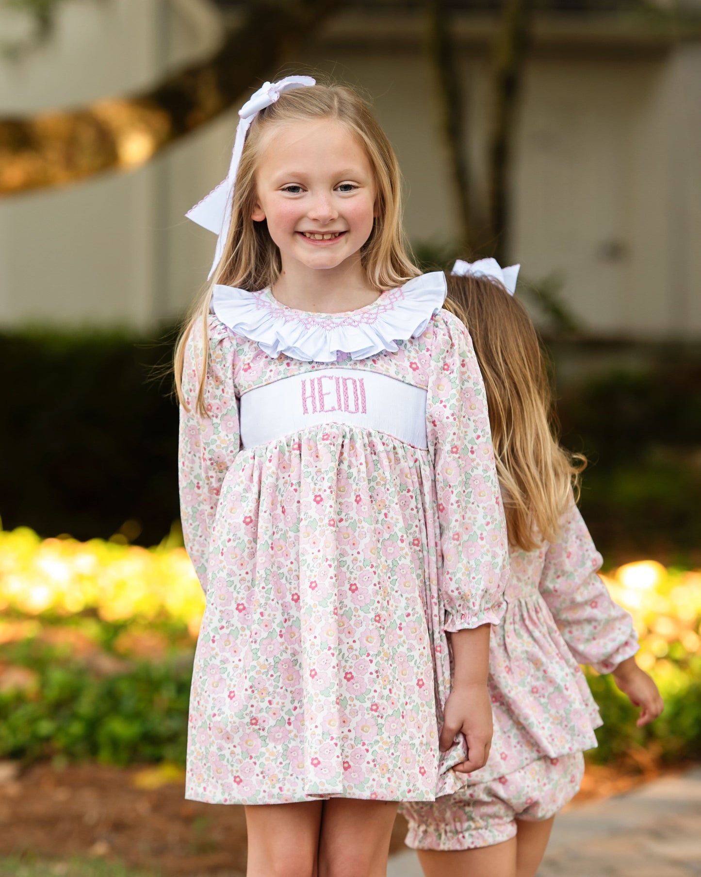 Young girl wearing a floral dress with a white ruffled collar, standing outdoors.