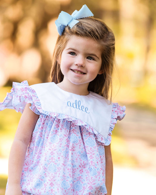 Young girl wearing a floral dress with 'adlee' embroidered on it, standing outdoors with a blurred natural background.