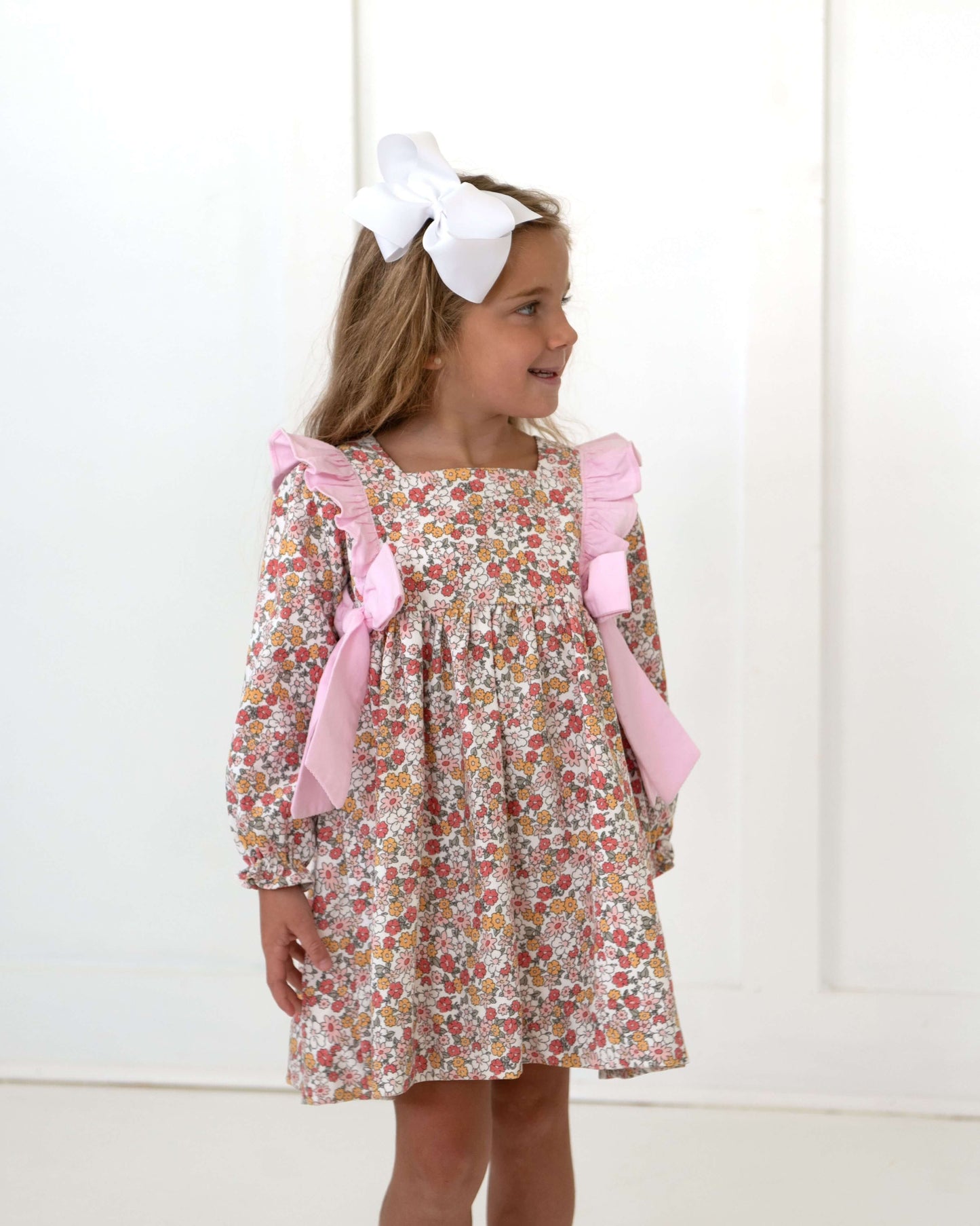 Young girl wearing a floral dress with pink bows and a large white bow in her hair against a white background.