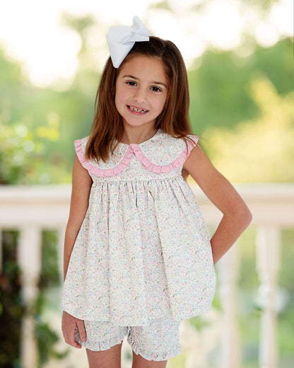 Young girl wearing a floral dress with pink trim and a white bow in her hair, standing outdoors.