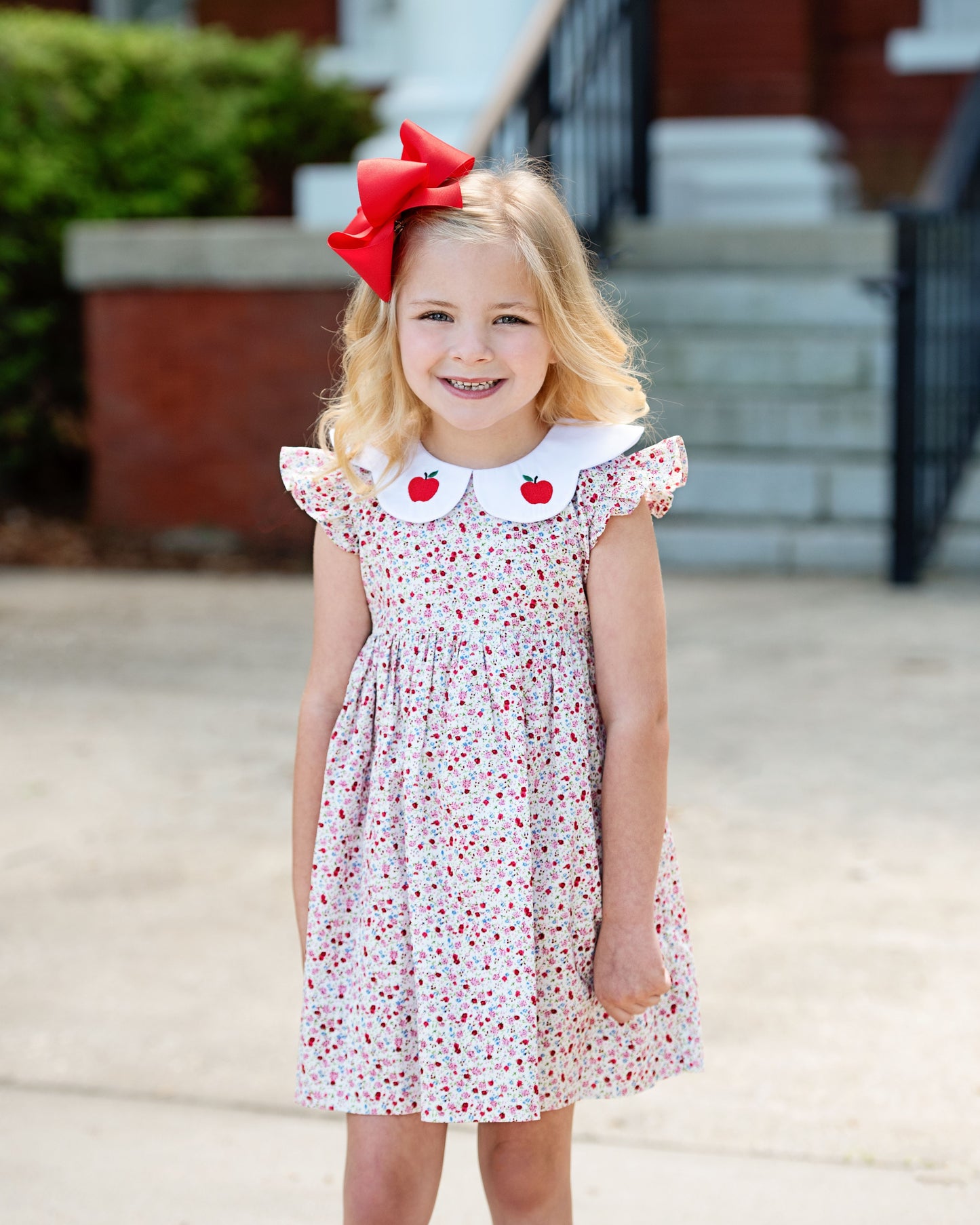 Young girl wearing a floral dress with red apple details and a red bow, standing outdoors.