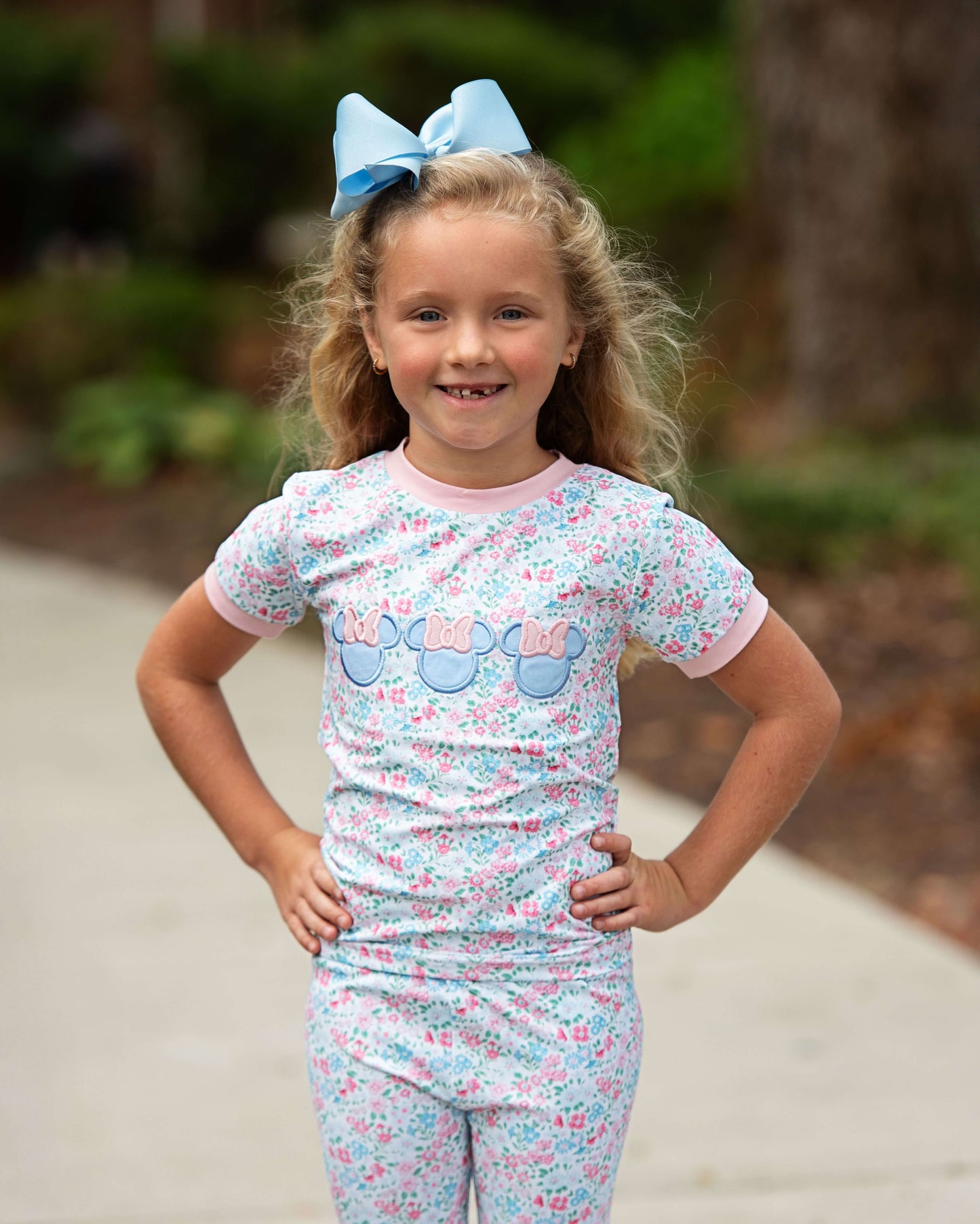 Young girl wearing a floral pajama set with a blue bow in her hair, standing outdoors.