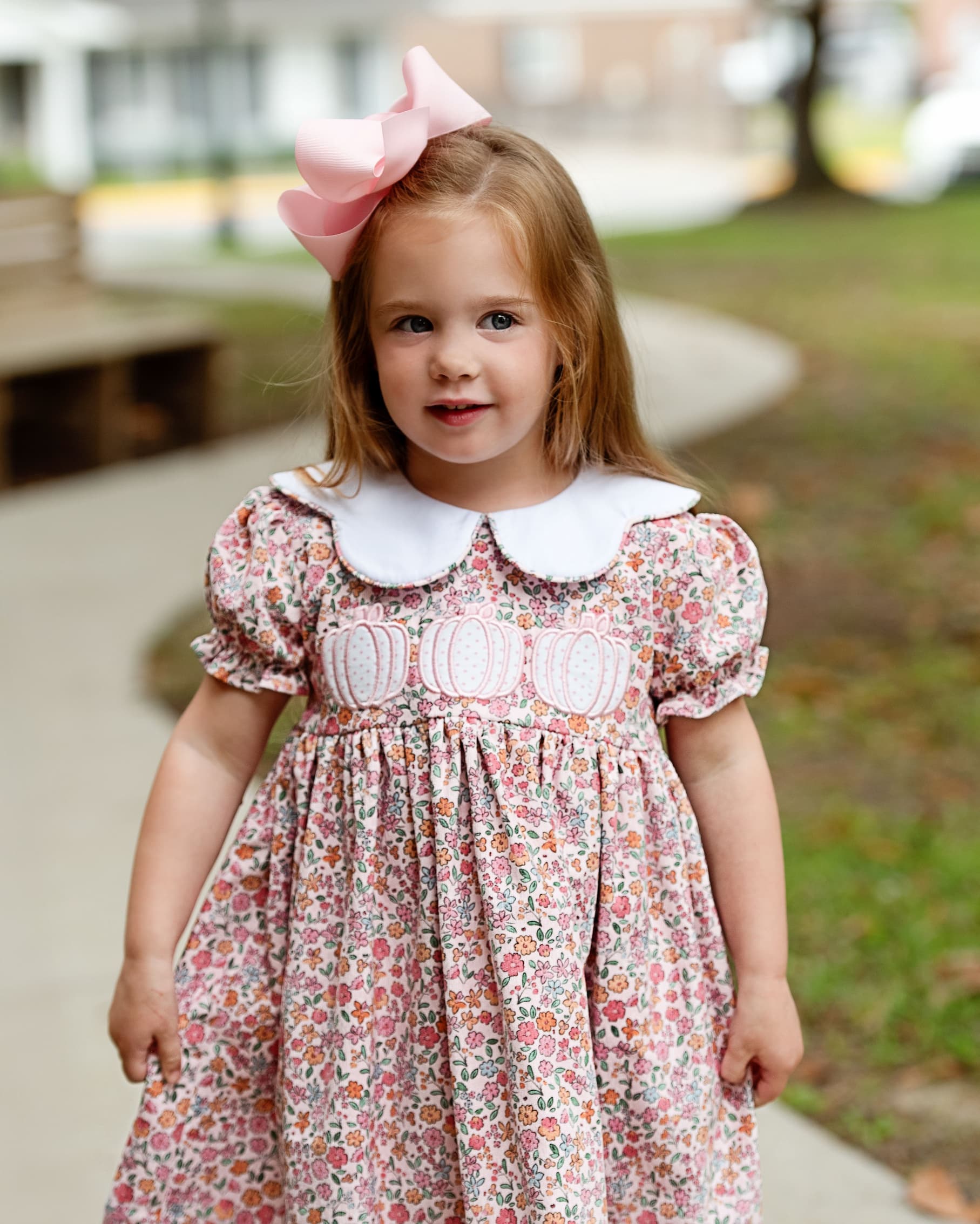 Young girl wearing a floral pumpkin dress with a white collar and pink bow outdoors.