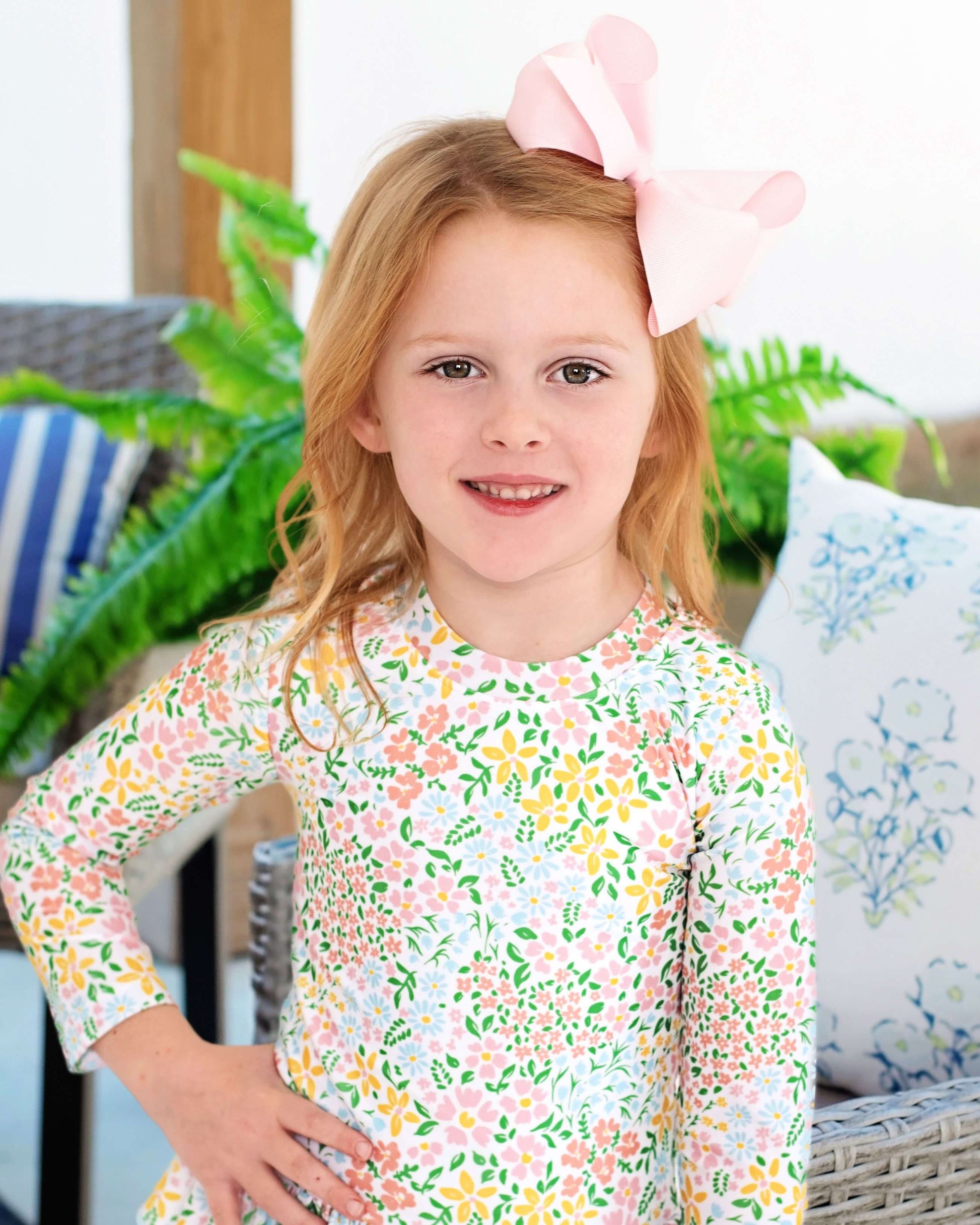 Young girl wearing a floral rashguard with a pink bow in her hair, sitting on a couch with plants in the background.