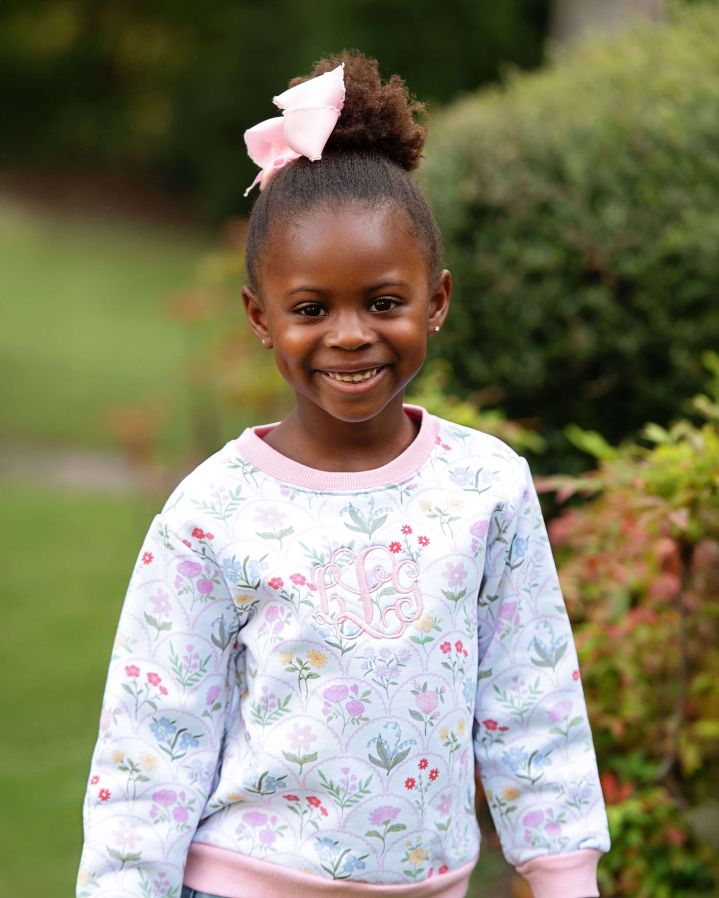 Young girl wearing a floral sweatshirt with a pink bow in her hair, standing outdoors.