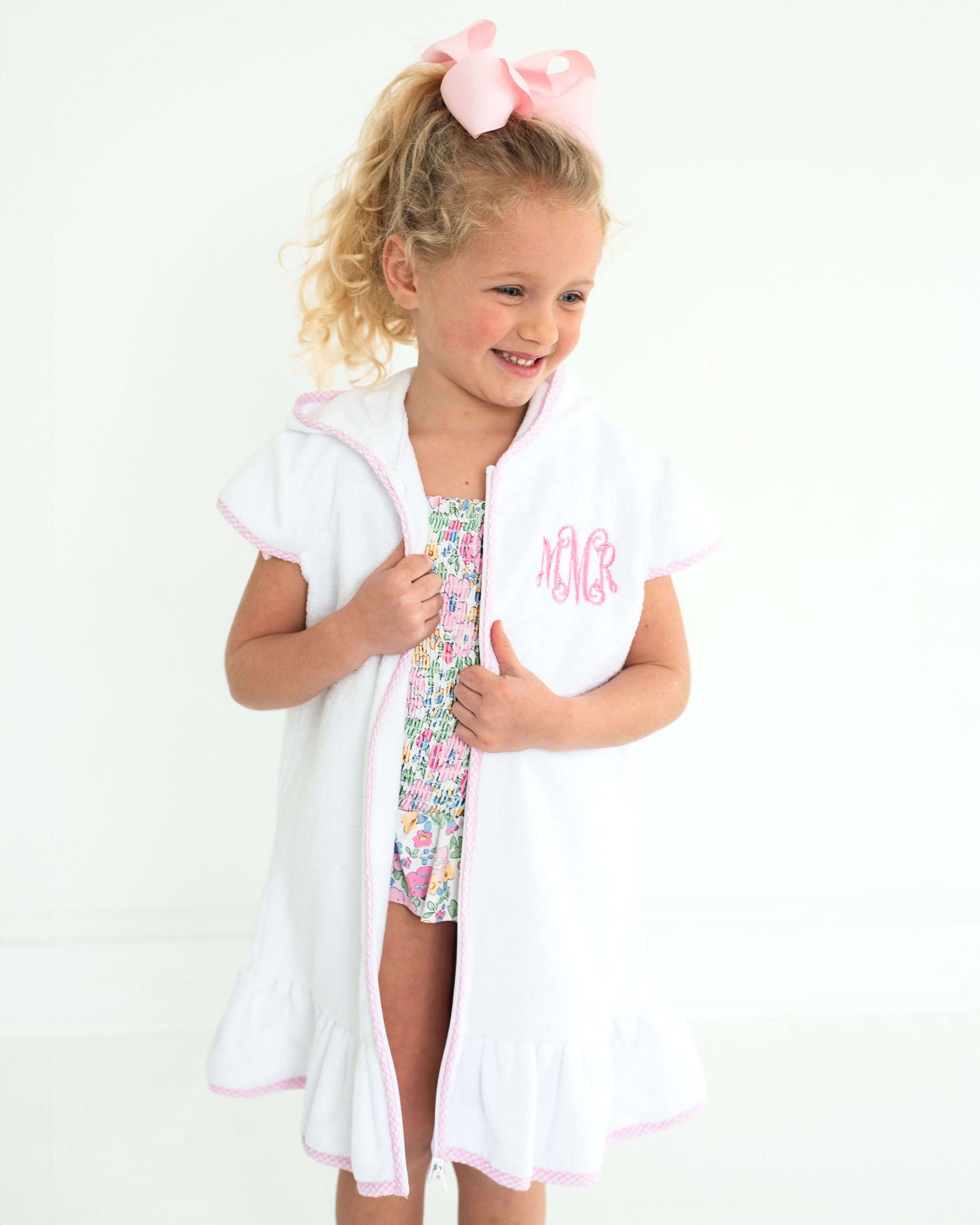 Young girl wearing a floral swimsuit with a white cover-up and pink hair bow on a white background