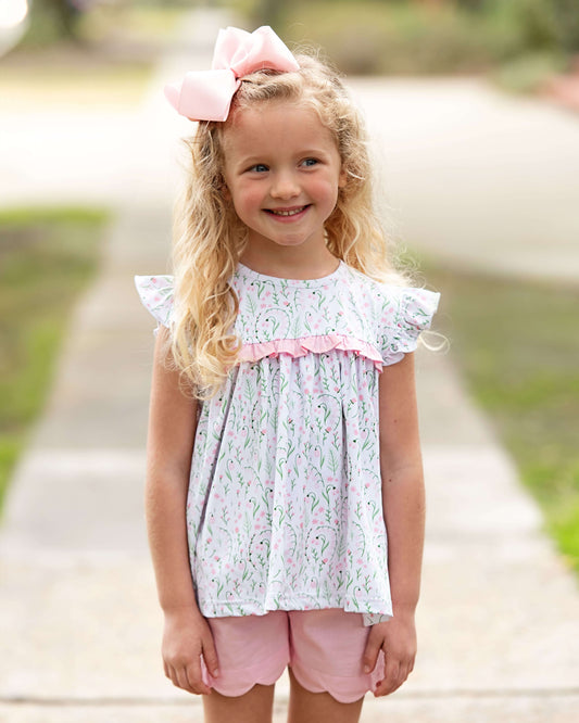 Young girl wearing a floral top and pink shorts with a bow in her hair, standing outdoors.