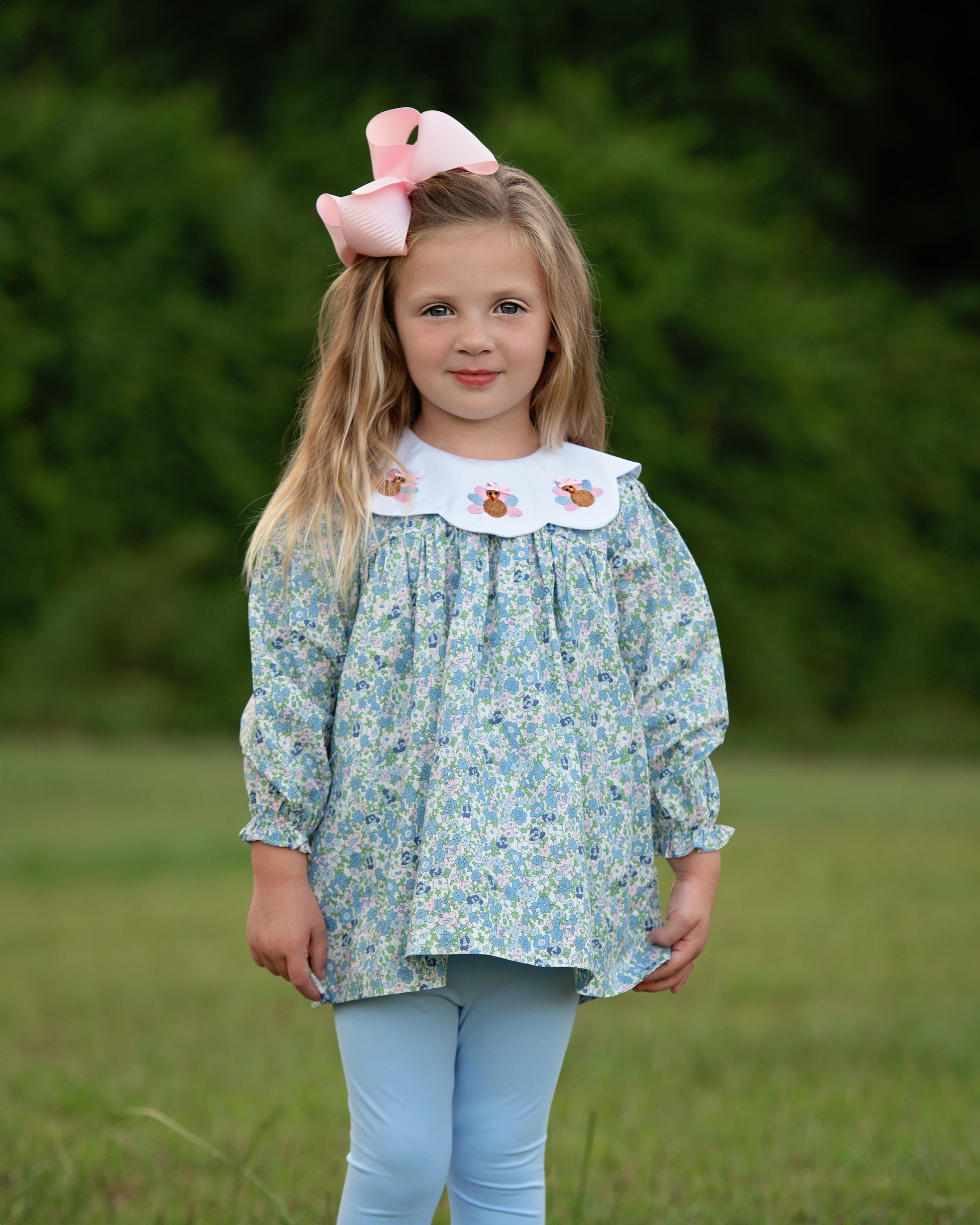 Young girl wearing a floral top with smocked turkeys legging set with a large pink bow outdoors