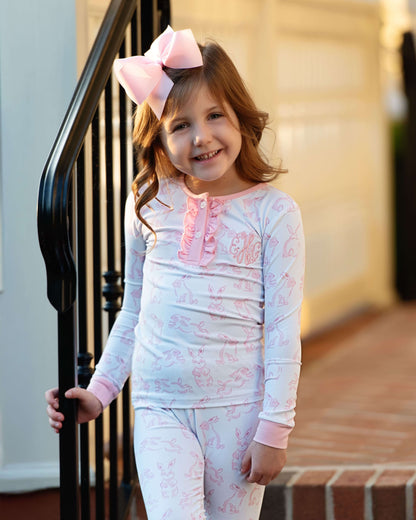 Young girl wearing a light blue and pink pajama set with a large bow in her hair, standing on a staircase.