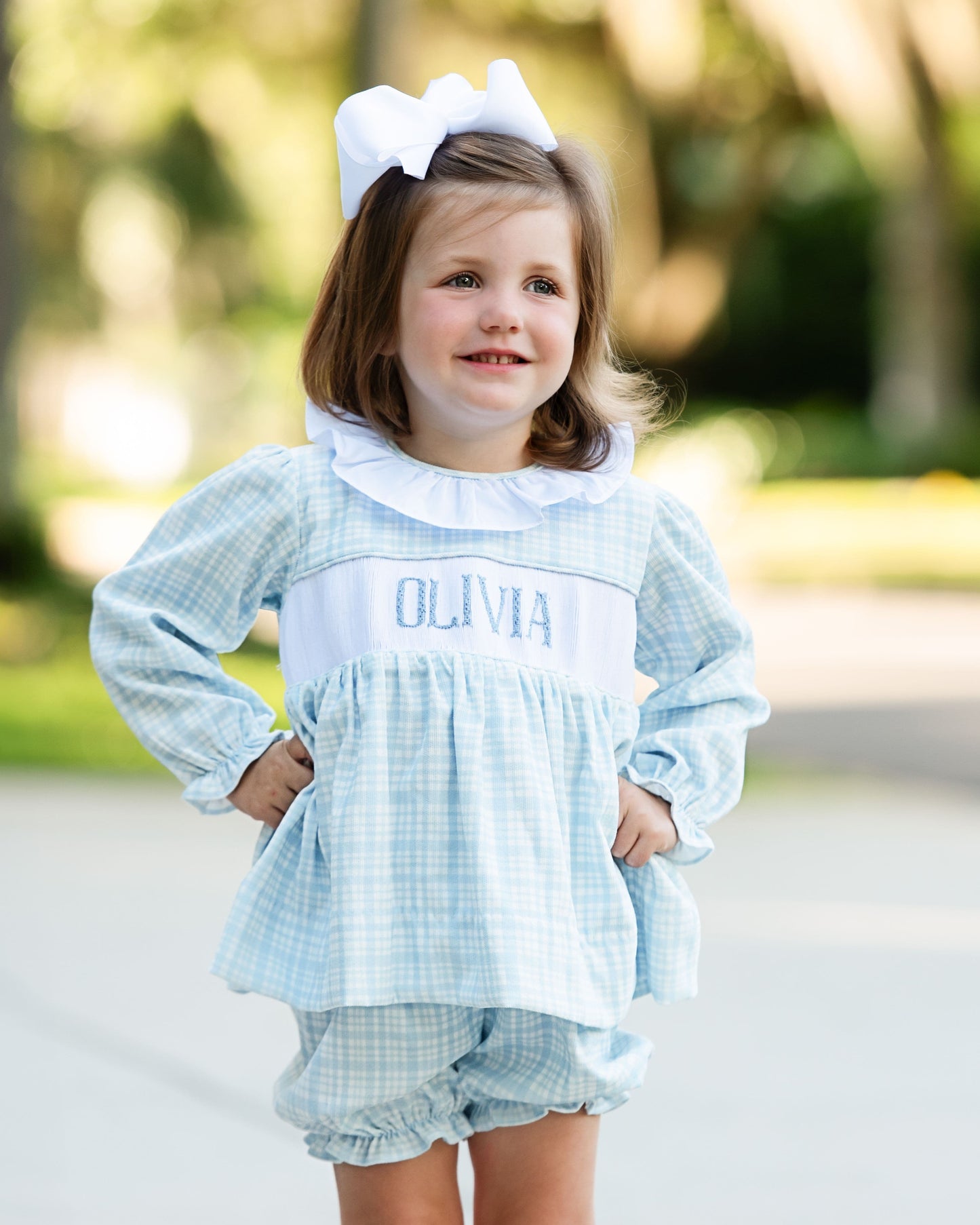 Young girl wearing a light blue checkered outfit with 'OLIVIA' printed on it, standing outdoors.