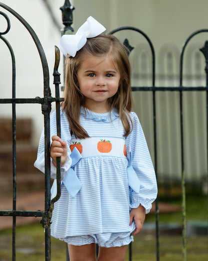Young girl wearing a light blue diaper set with pumpkin design, standing in front of a black metal gate.