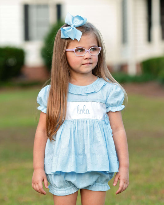 Young girl wearing a light blue dress with 'hello' on it, standing outdoors.