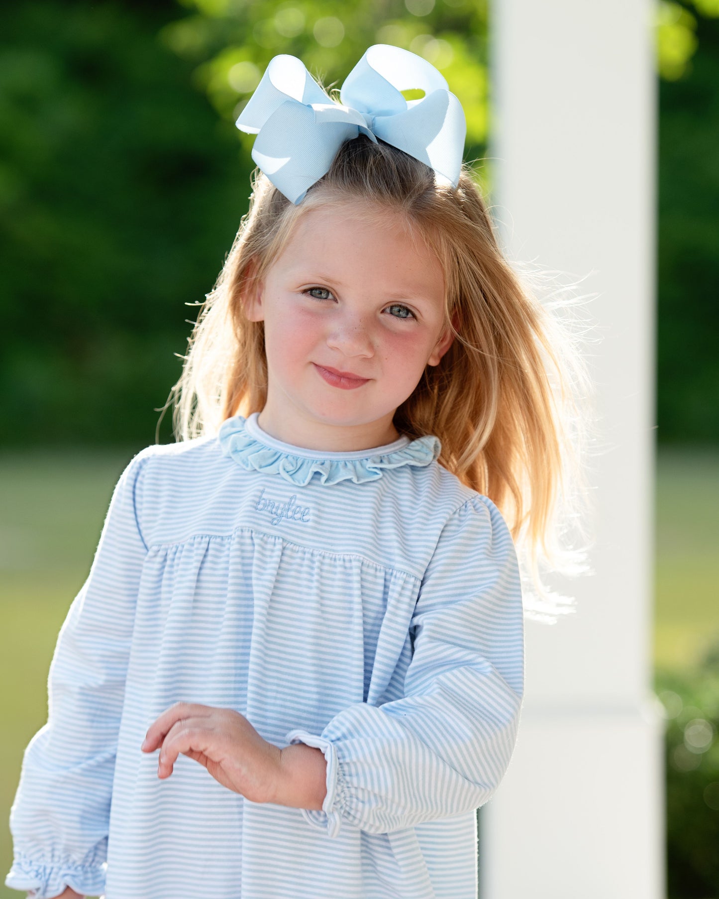 Young girl wearing a light blue dress with a large bow in her hair, standing outdoors.