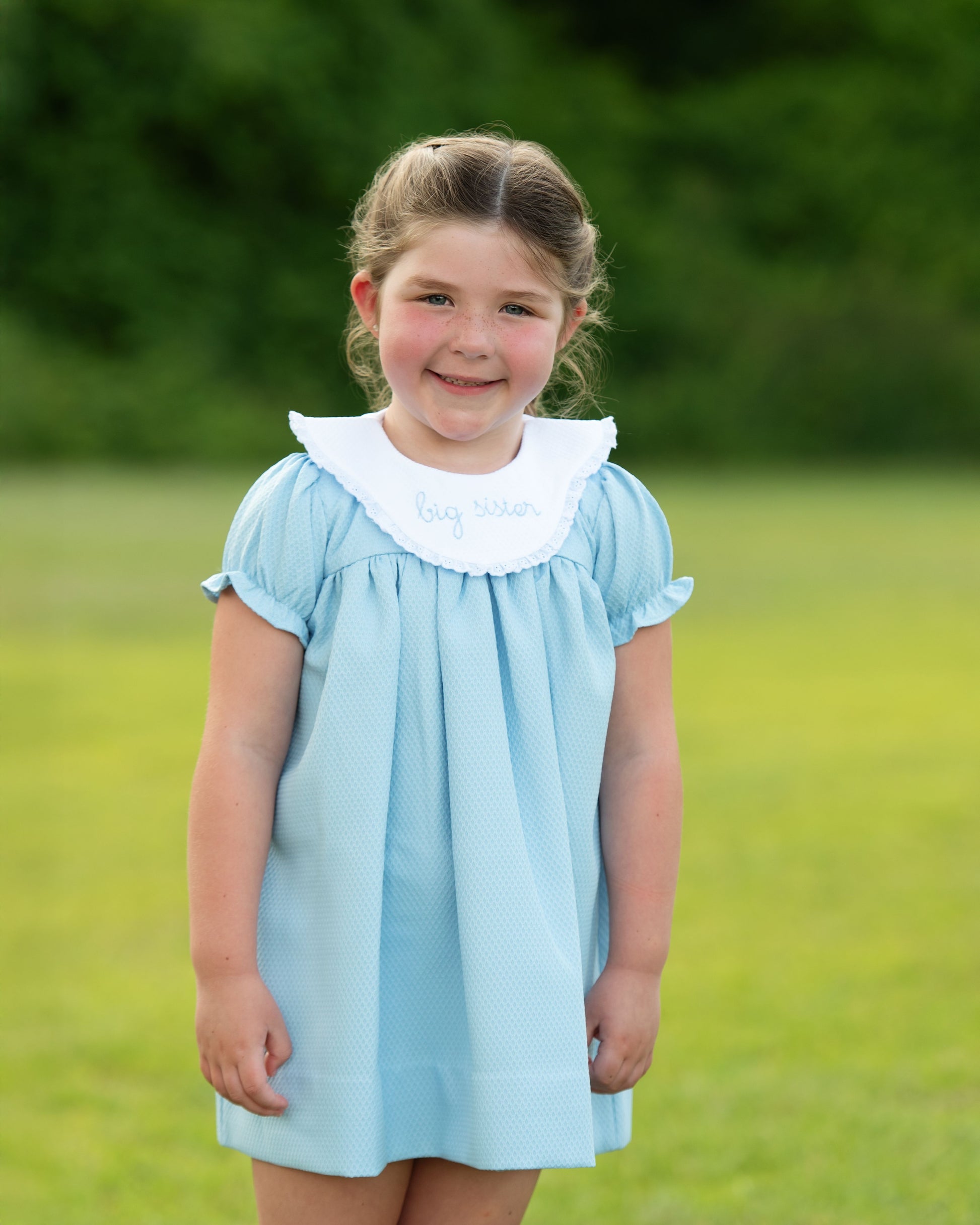 Young girl wearing a light blue dress with a white collar in an outdoor setting.