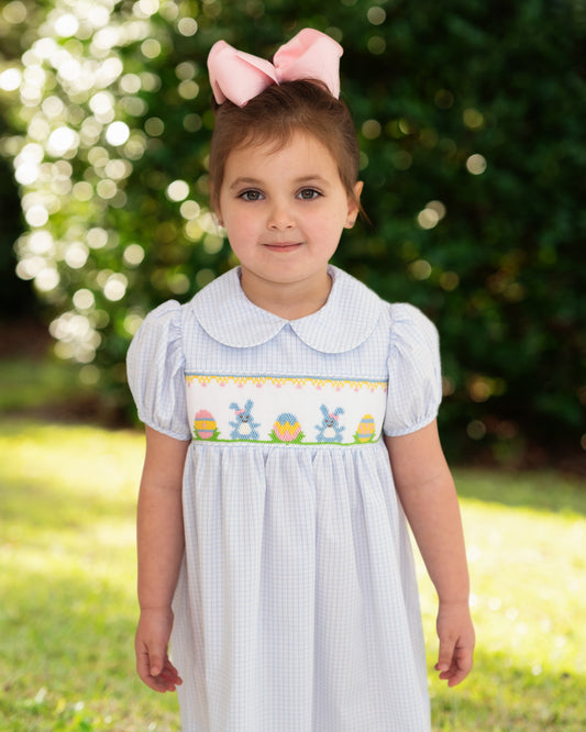 Young girl wearing a light blue dress with bunny and Easter egg design outdoors.
