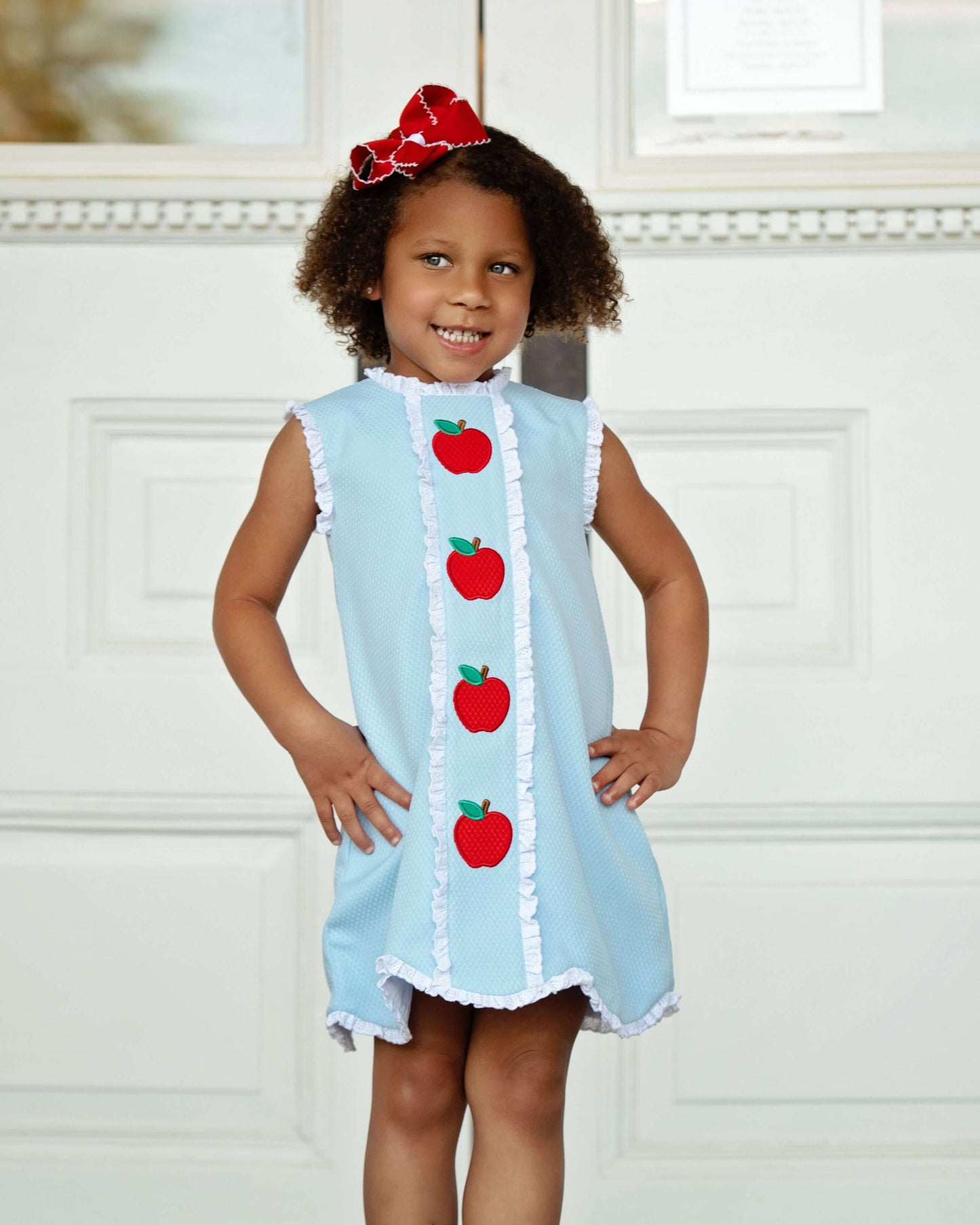 Young girl wearing a light blue dress with red apple patterns against a white background