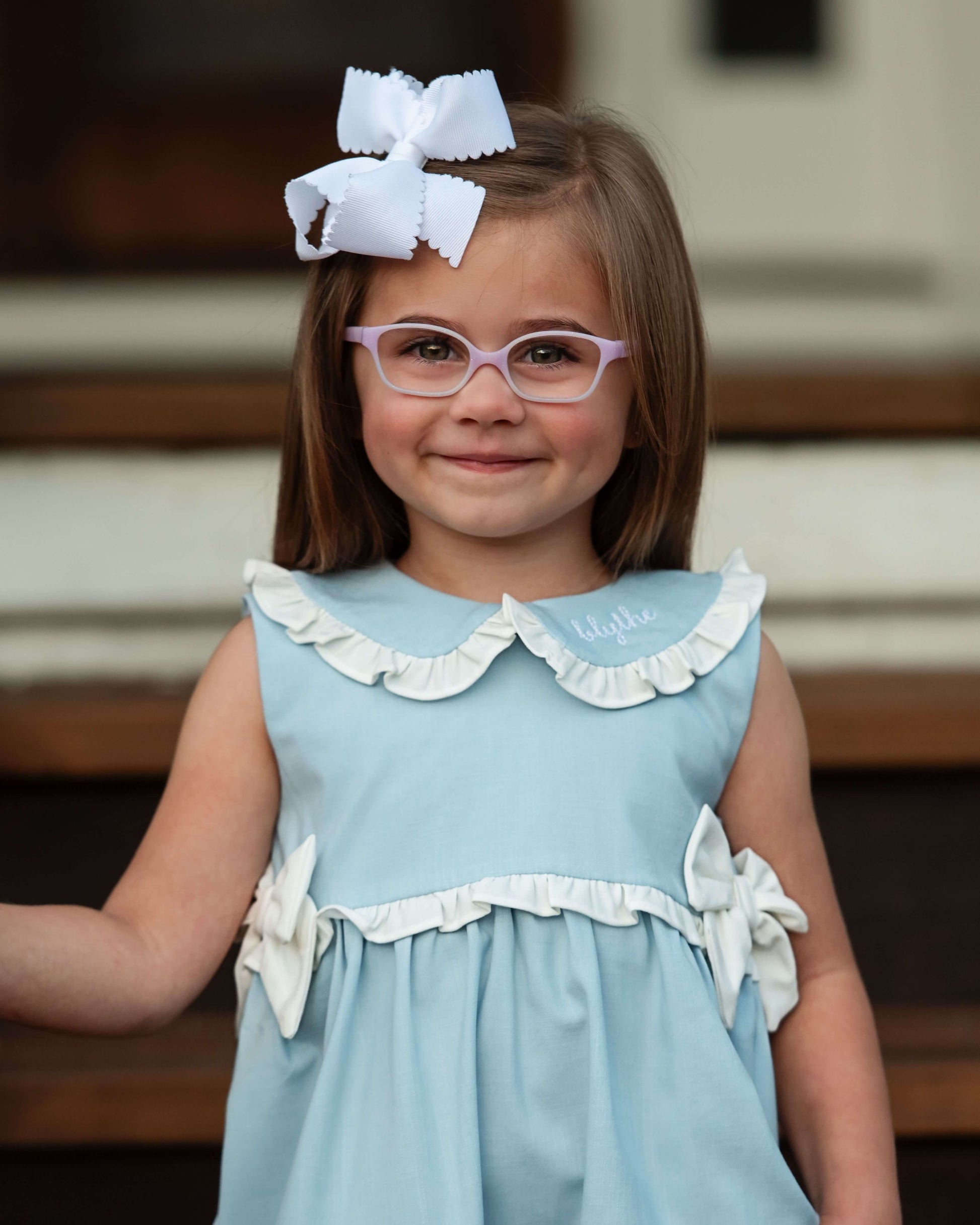 Young girl wearing a light blue dress with white ruffles and a large white bow in her hair.