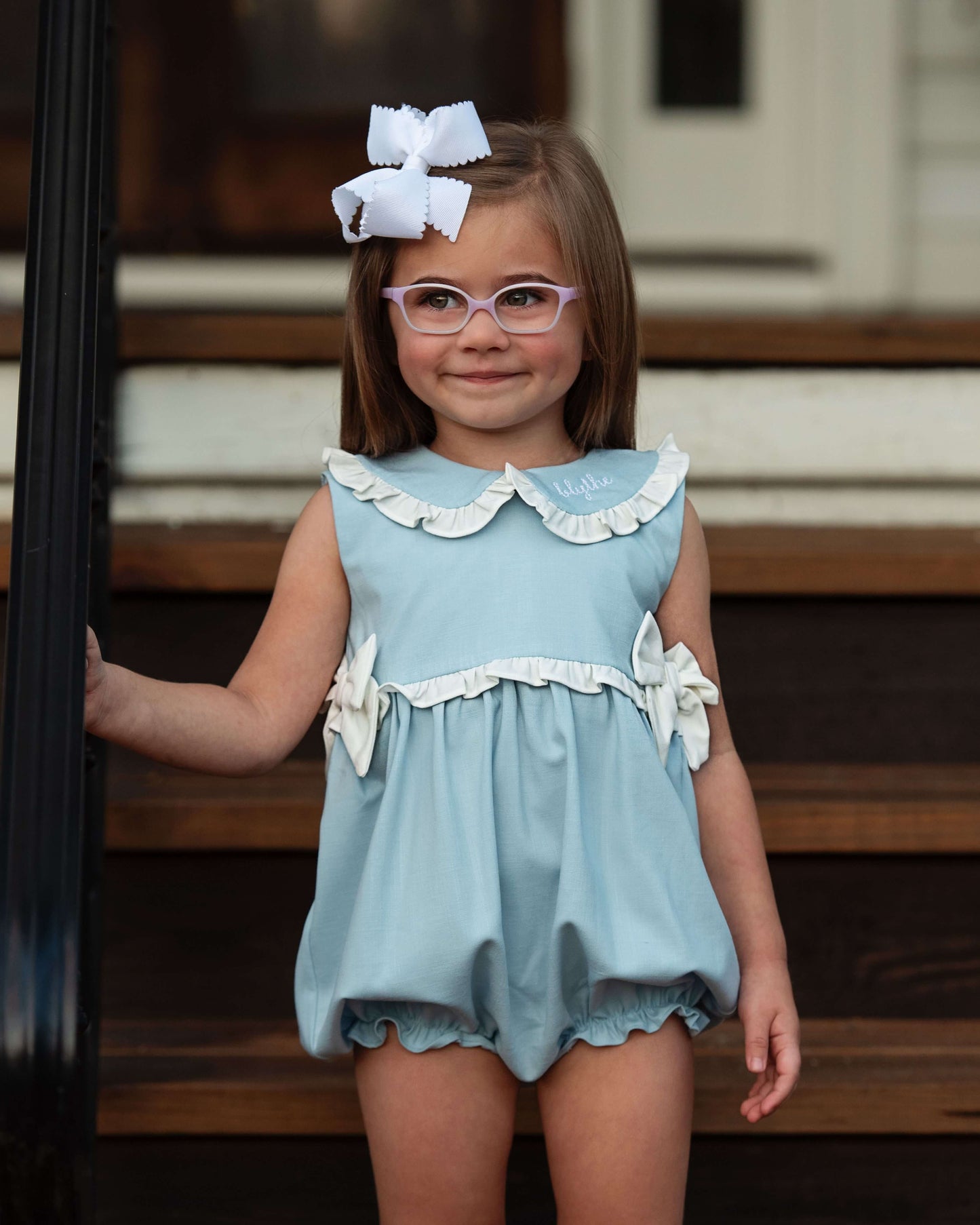 Young girl wearing a light blue dress with white ruffles and bows, standing on wooden steps.