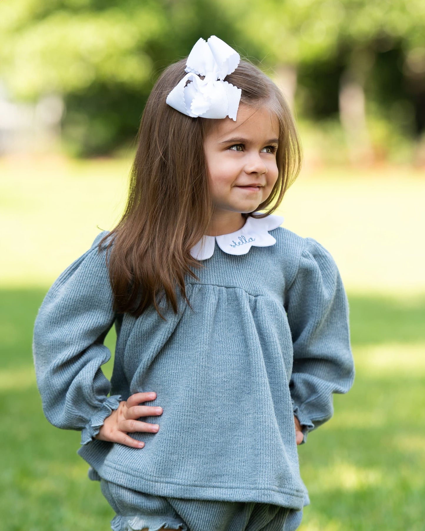 Young girl wearing a light blue outfit with a white collar and large white bow outdoors.