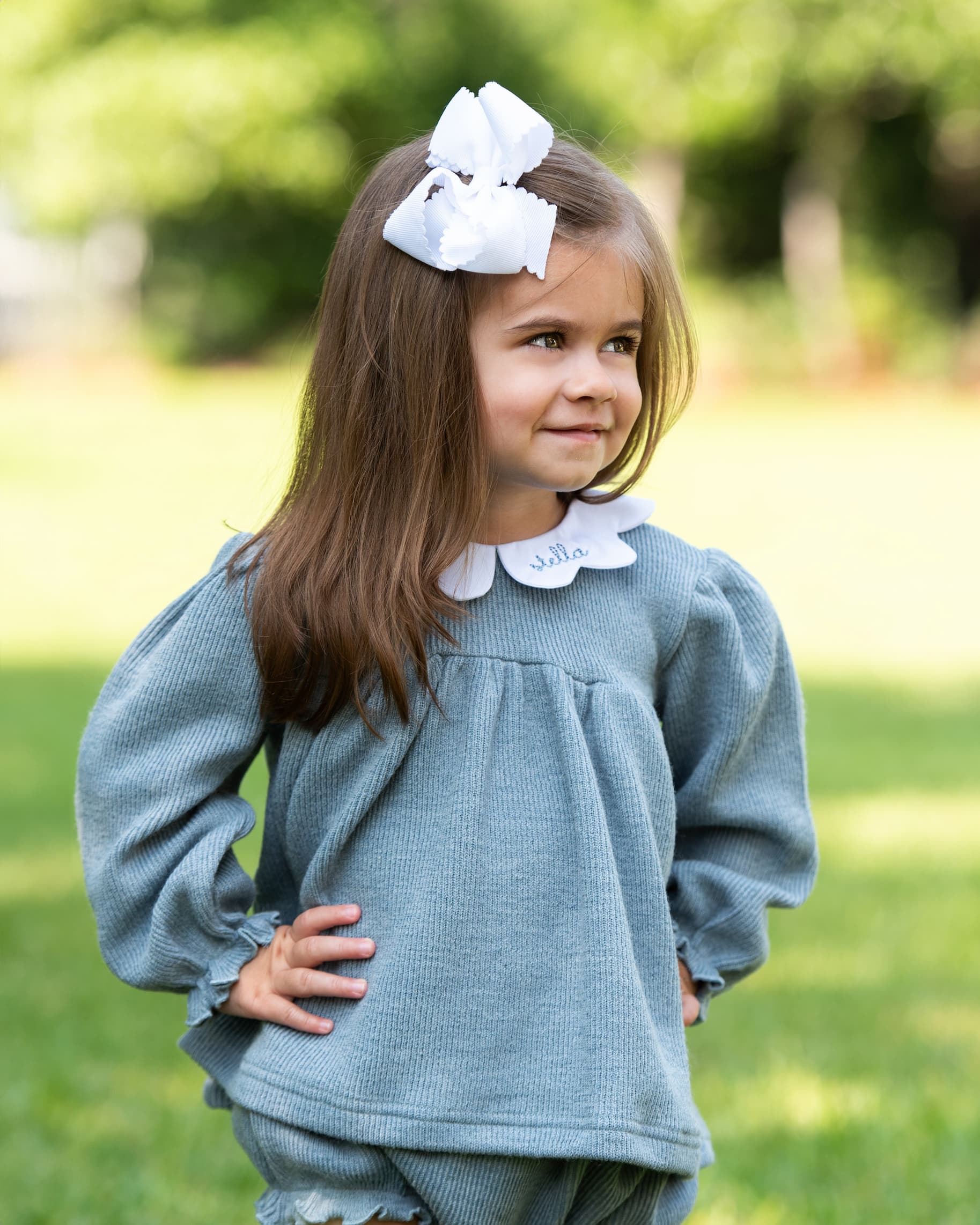 Young girl wearing a light blue outfit with a white collar and large white bow outdoors.