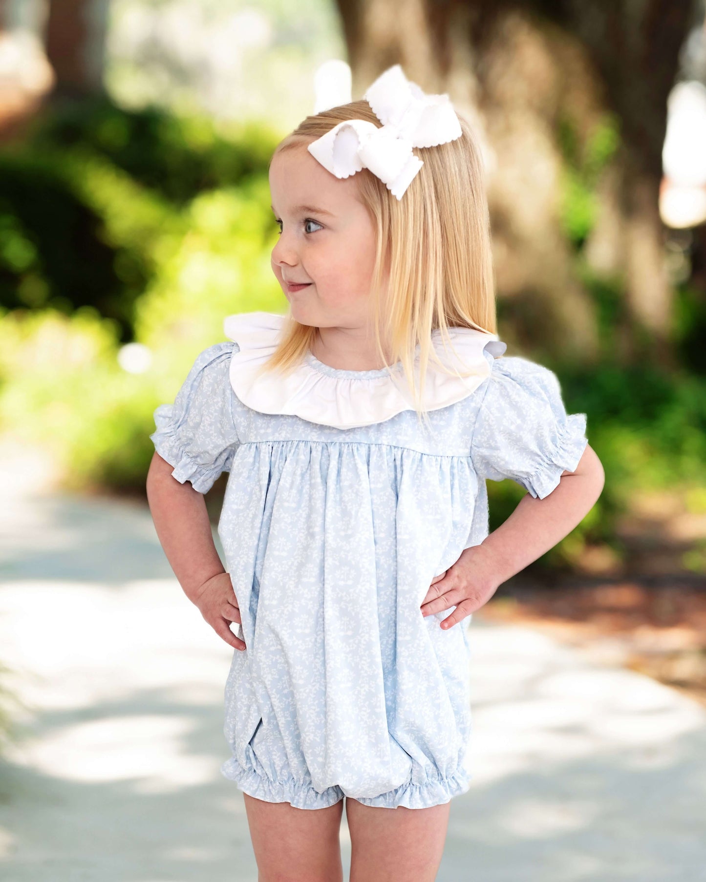 Young girl wearing a light blue romper with a white bow in her hair, standing outdoors.