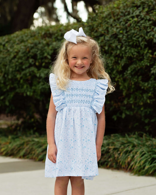 Young girl wearing a light blue smocked dress with ruffled sleeves in front of green bushes.