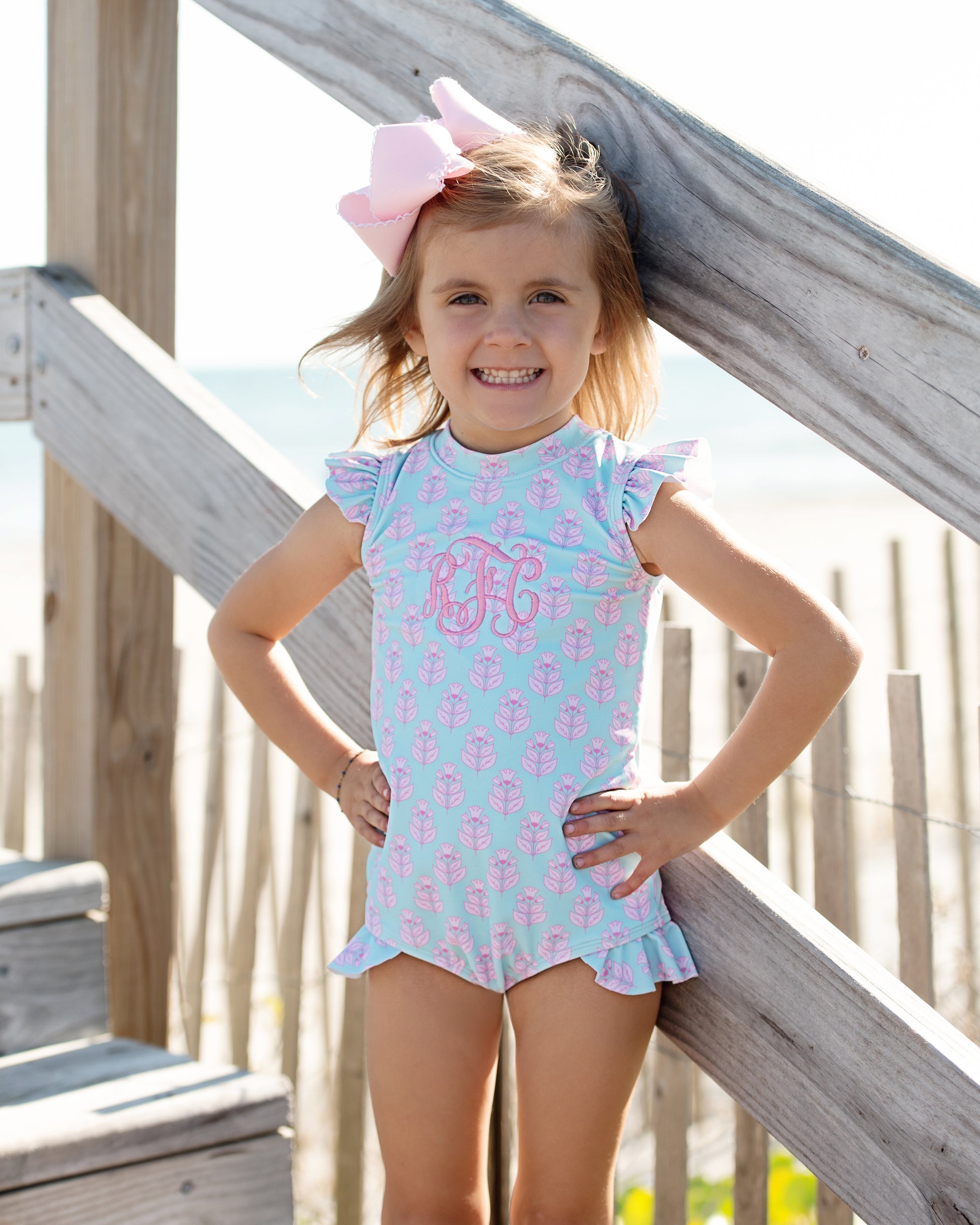 Young girl wearing a light blue swimsuit with pink anchors and a large pink bow, standing on wooden steps.