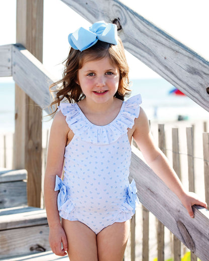 Young girl wearing a light blue swimsuit with ruffles and a large bow, standing on a wooden deck.