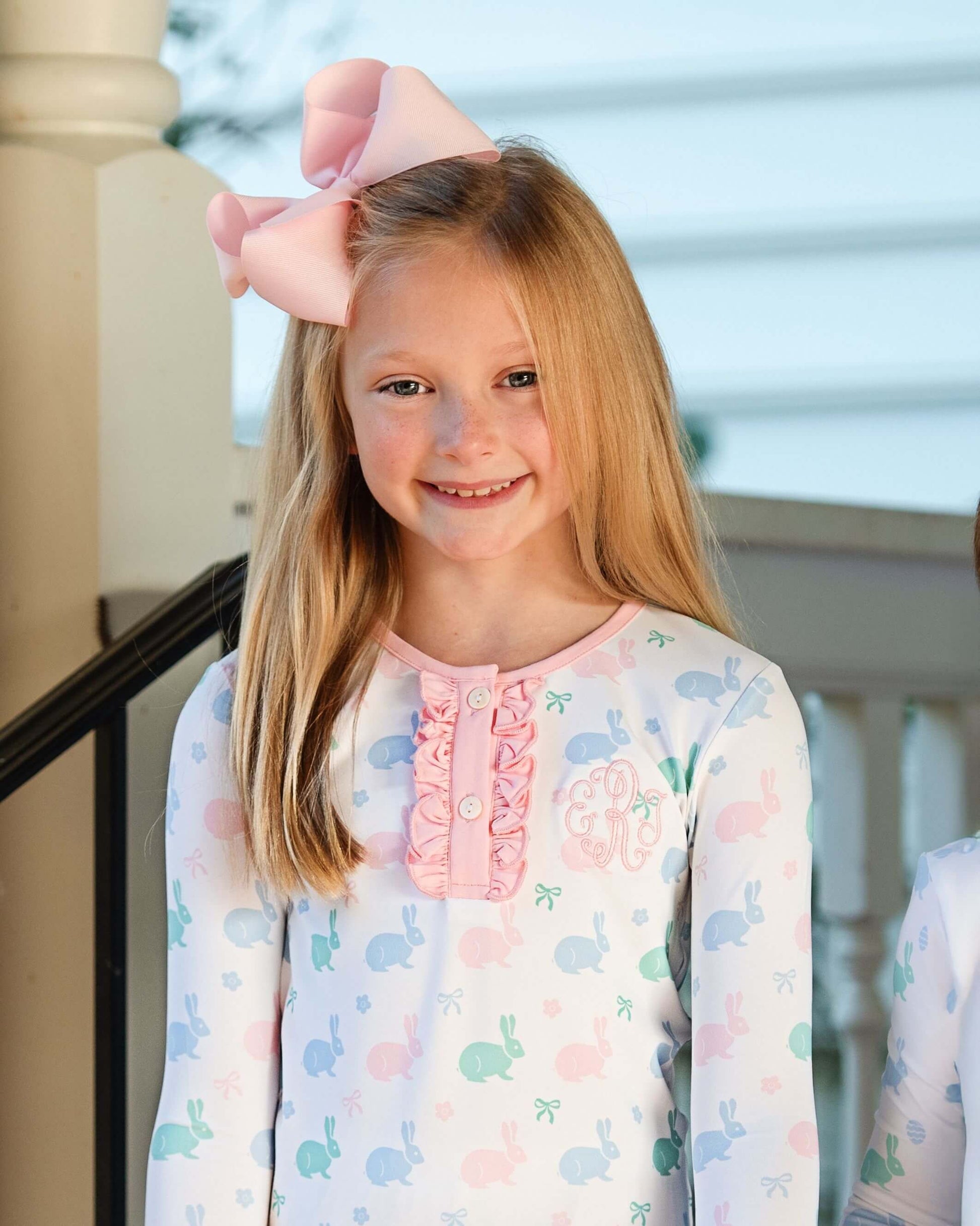 Young girl wearing a pastel floral dress with a large pink bow on her head, standing outdoors.