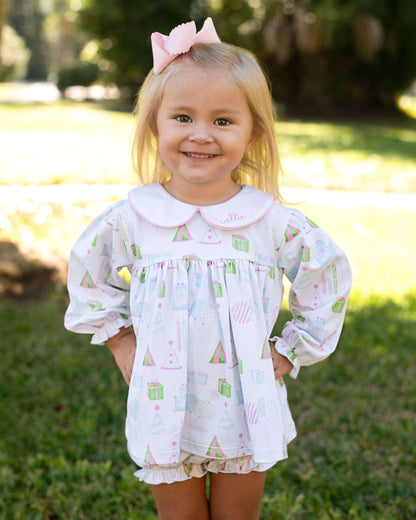 Young girl wearing a patterned blouse and shorts with a pink bow in her hair, standing outdoors.
