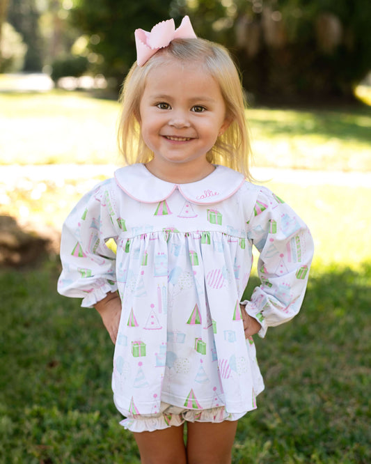 Young girl wearing a patterned blouse and shorts with a pink bow in her hair, standing outdoors.
