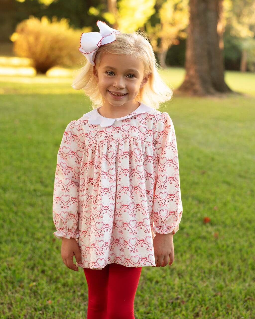 Young girl wearing a patterned dress and red leggings outdoors on a grassy area.