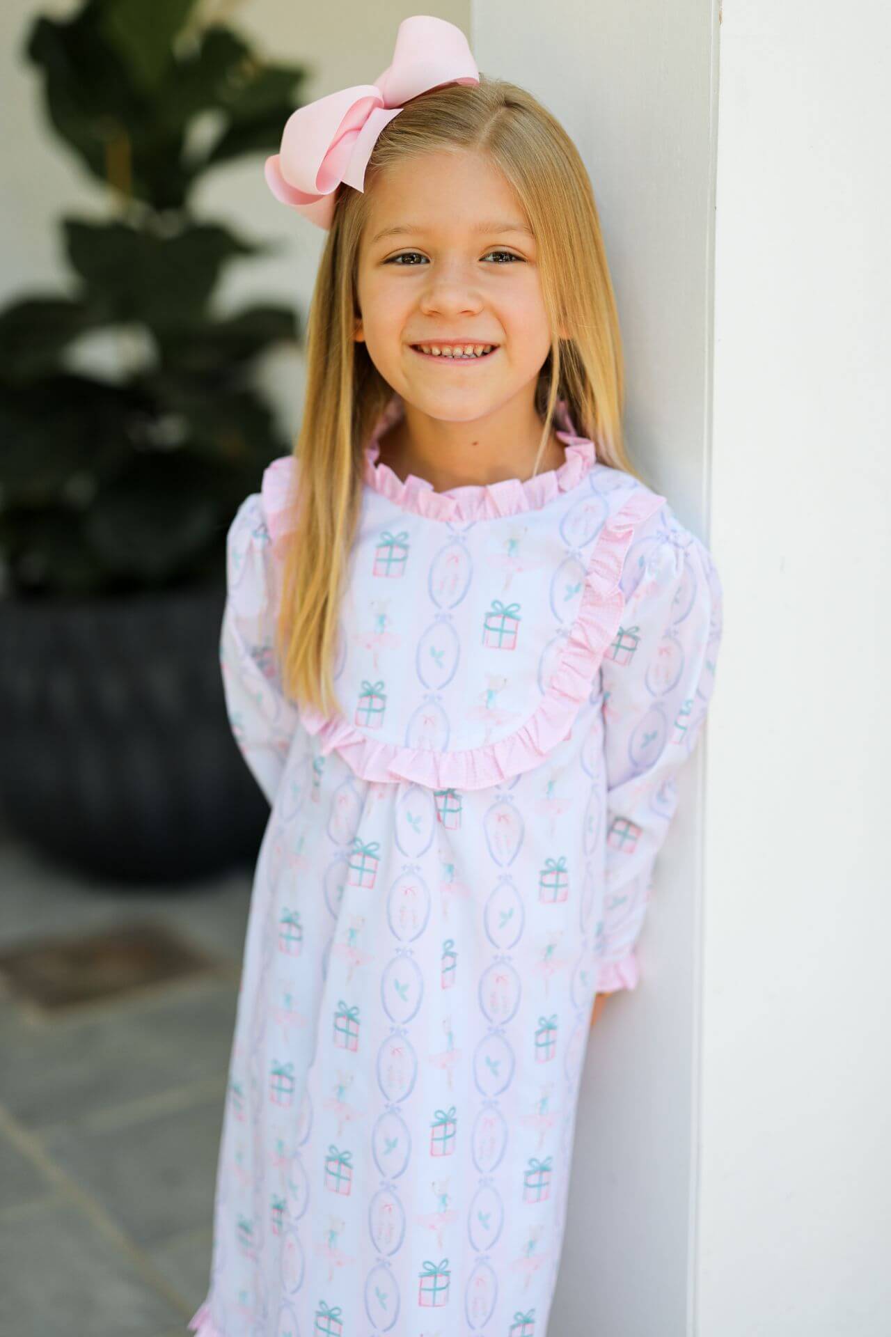 Young girl wearing a patterned holiday nightgown with a pink bow in her hair, standing against a white wall.