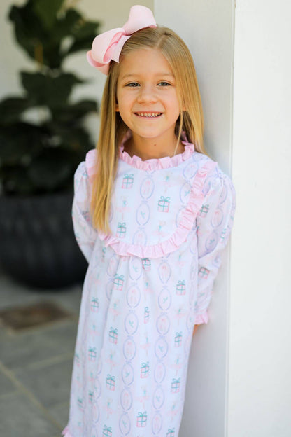 Young girl wearing a patterned holiday nightgown with a pink bow in her hair, standing against a white wall.