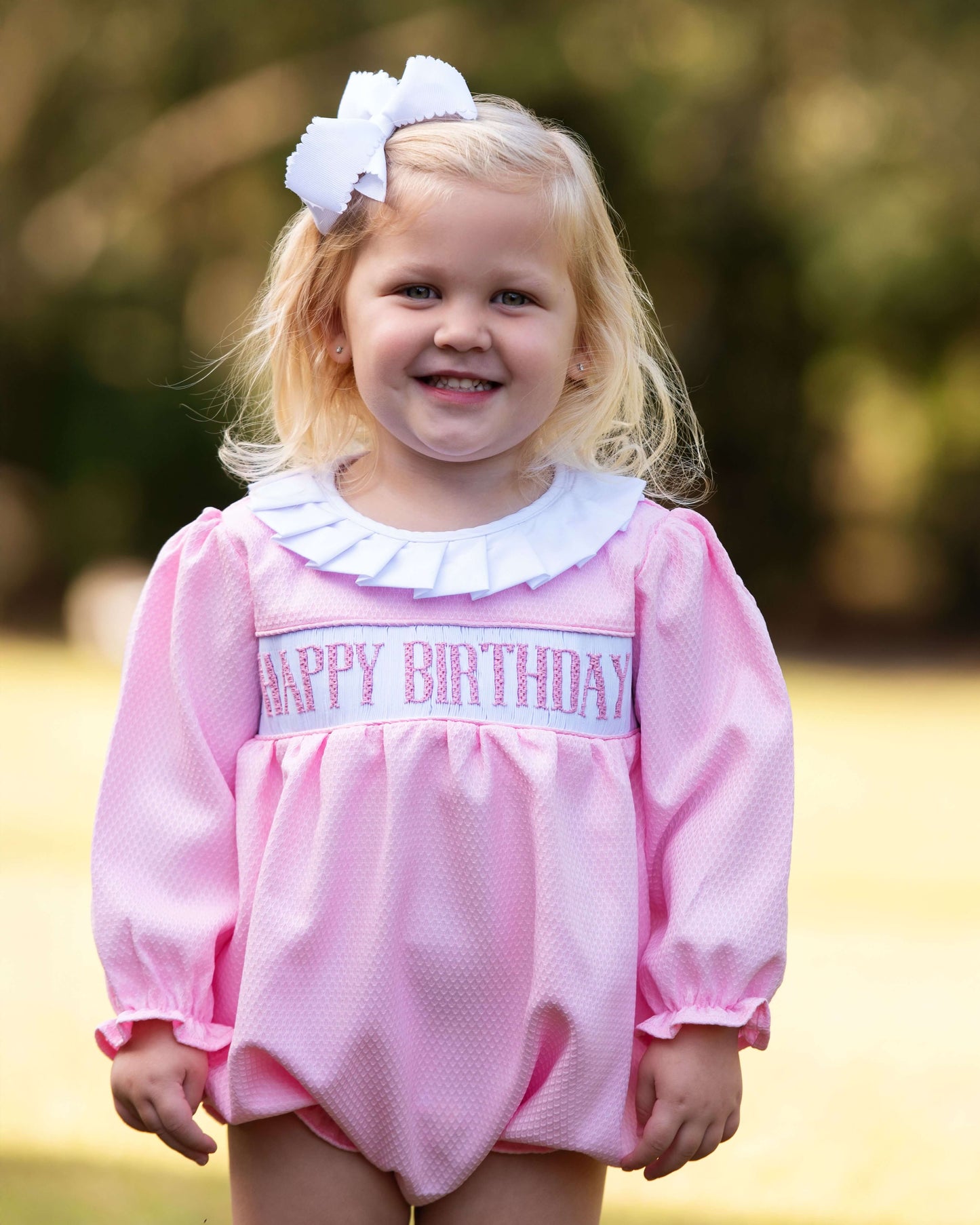 Young girl wearing a pink 'Happy Birthday' dress outdoors with a blurred background