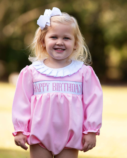 Young girl wearing a pink 'Happy Birthday' dress outdoors with a blurred background